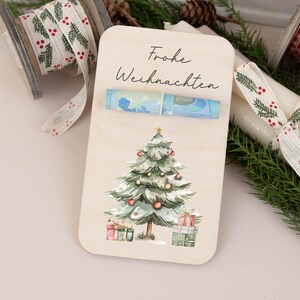 May include: A wooden card with the text "Frohe Weihnachten" and a watercolor illustration of a Christmas tree with presents underneath. The card has a slot for a rolled up bill.