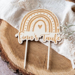 May include: Wooden rainbow cake topper with the text "Finn's Taufe". The rainbow design features layered arches and heart details. The topper is light brown and has two pointed sticks for insertion into a cake.