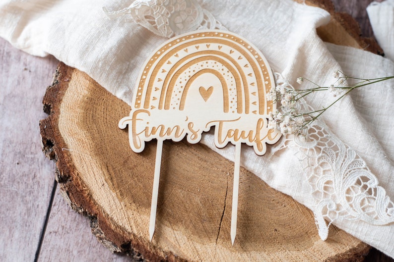 May include: Wooden rainbow cake topper with the text "Finn's Taufe". The rainbow design features layered arches and heart details. The topper is light brown and has two pointed sticks for insertion into a cake.