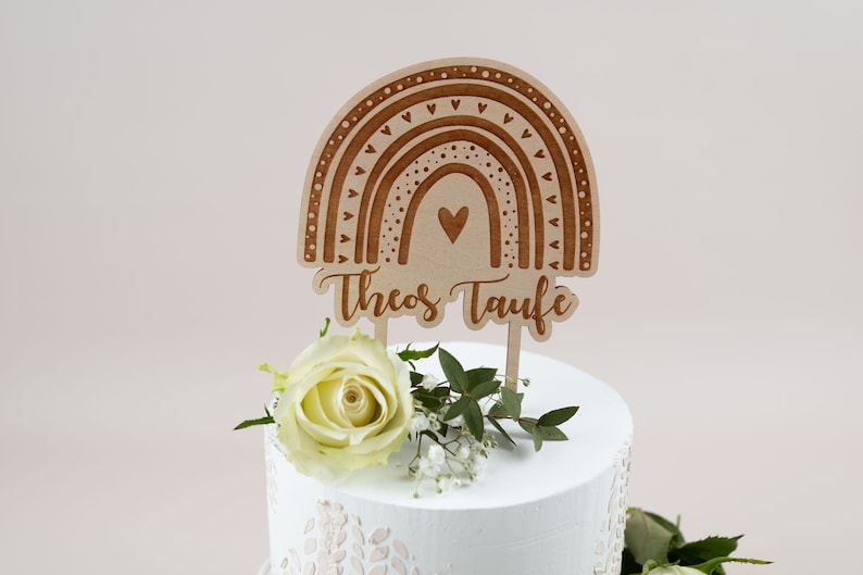 May include: Wooden cake topper with a rainbow design and the text "Theos Taufe". The topper is brown and features a heart in the center of the rainbow. A white rose and greenery are at the base of the cake.