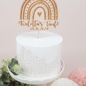 May include: A white cake with a decorative leaf pattern, topped with a wooden rainbow cake topper. The topper has the text "Kalotta's Taufe" and the date "26.10.2024". The cake sits on a white cake stand.