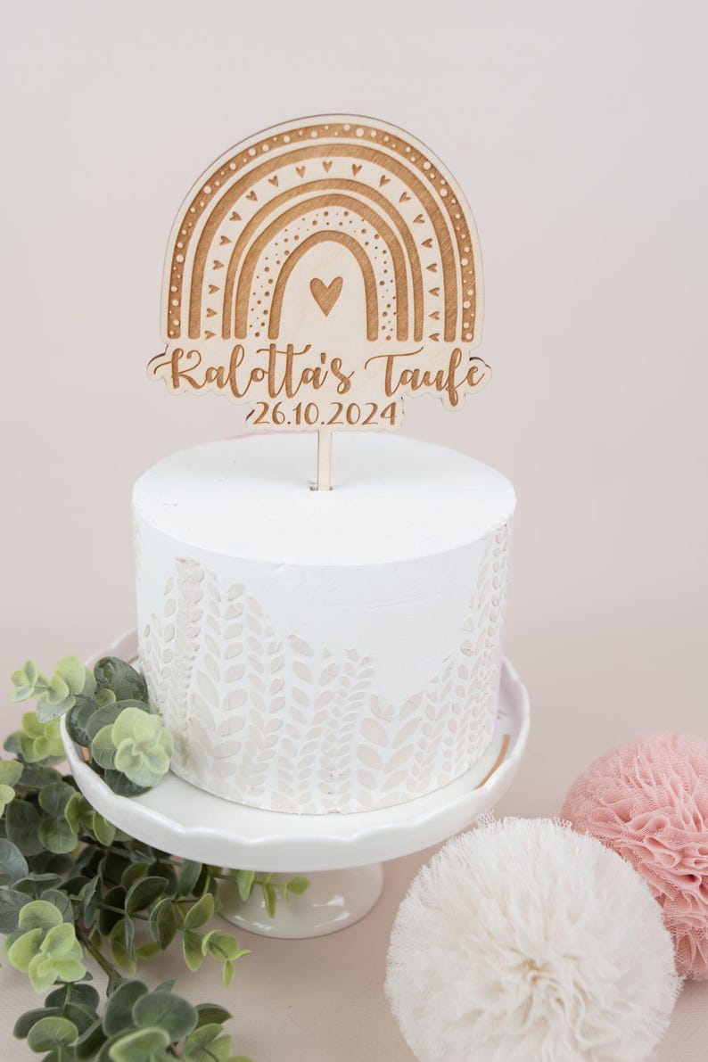 May include: A white cake with a decorative leaf pattern, topped with a wooden rainbow cake topper. The topper has the text "Kalotta's Taufe" and the date "26.10.2024". The cake sits on a white cake stand.