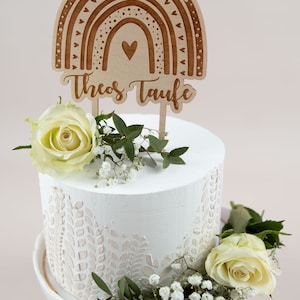 May include: A white cake with floral decorations and a wooden rainbow cake topper that reads "Theos Taufe". The cake is adorned with white roses and greenery, set on a white cake stand.