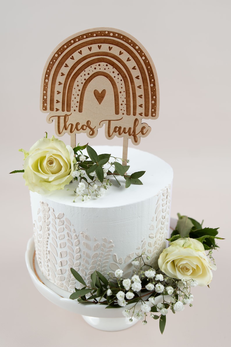 May include: A white cake with floral decorations and a wooden rainbow cake topper that reads "Theos Taufe". The cake is adorned with white roses and greenery, set on a white cake stand.