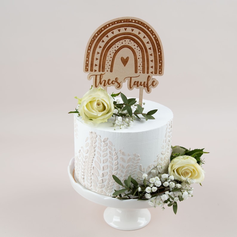 May include: A white cake with a decorative floral design and a wooden rainbow cake topper that reads "Theos Taufe". The cake is decorated with white roses and greenery, and sits on a white cake stand.