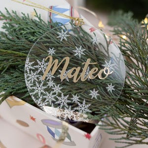 Könnte beinhalten: Ein runder, transparenter Weihnachtsbaumschmuck mit einem goldenen Glitzer-Schneeflockenmuster und dem Namen "Mateo" in goldenen Buchstaben.