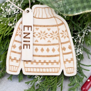 Peut inclure: Une d&eacute;coration de No&euml;l en bois en forme de pull avec un motif brun et blanc. L'ornement a une &eacute;tiquette attach&eacute;e qui dit "ENIE".