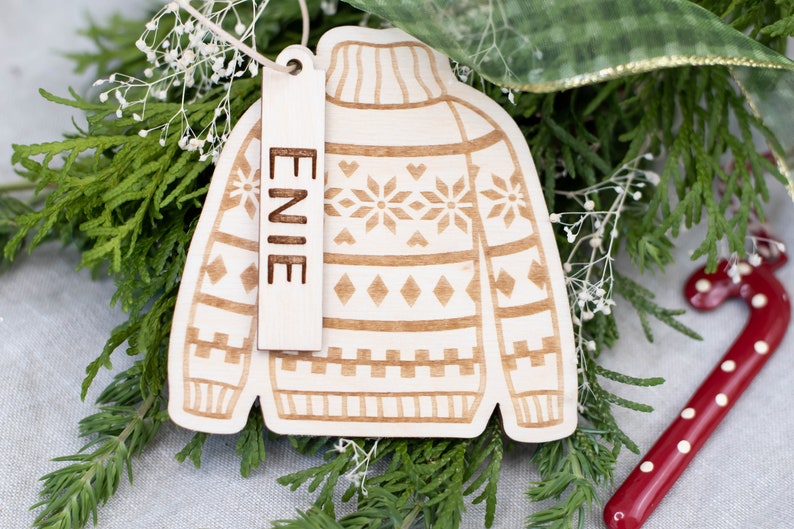 Peut inclure: Une d&eacute;coration de No&euml;l en bois en forme de pull avec un motif brun et blanc. L'ornement a une &eacute;tiquette attach&eacute;e qui dit "ENIE".
