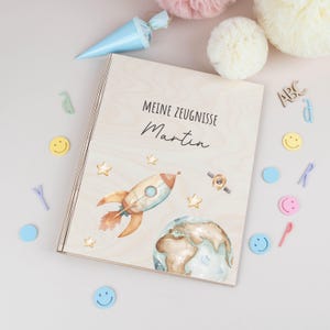 Puede incluir: Una caja de madera con el texto "Meine Zeugnisse Martin" y una ilustración en acuarela de un cohete, estrellas y la Tierra. La caja está rodeada de elementos decorativos como emoticonos y letras.