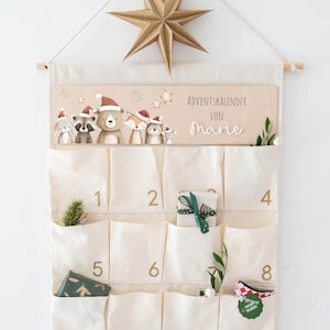Può includere: Un calendario dell'avvento in tessuto color crema appeso a un'asta di legno con una stella dorata. Il pannello in legno presenta illustrazioni di animali del bosco con cappelli di Babbo Natale e il testo "ADVENTSKALENDER VON Marie". Ha tasche numerate.