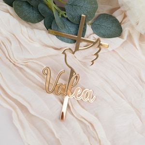 Può includere: Un cake topper dorato con una croce, una colomba e il nome "Valea" in corsivo. Il topper è posto su un tessuto color crema con foglie verdi sullo sfondo. Ideale per un battesimo o una cresima.
