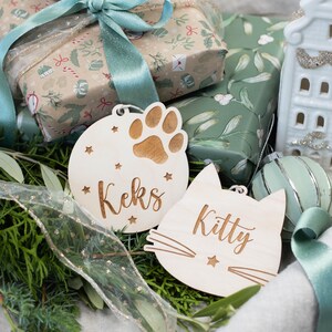 May include: Two wooden Christmas ornaments with personalized names. One ornament is round with a paw print and the name "Keks" and the other is shaped like a cat with the name "Kitty".