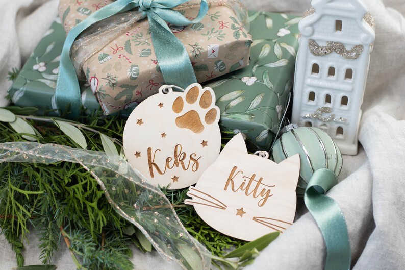 May include: Two wooden Christmas ornaments with personalized names. One ornament is round with a paw print and the name "Keks" and the other is shaped like a cat with the name "Kitty".