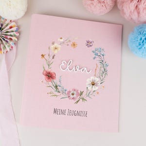 Op de afbeelding: Een lichtroze rechthoekige boekomslag met een centrale bloemenkrans in roze, blauwe, gele en groene tinten. De naam Elva staat in wit schrift in het midden, met de tekst MEINE ZEUGNISSE eronder gedrukt in zwarte letters.