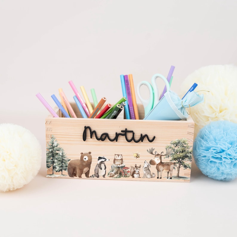 May include: A wooden pencil box with the name "Martin" in black script. The box is decorated with forest animals and filled with colorful pencils, pens, and scissors. Decorative paper pom-poms are in the background.