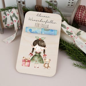 May include: A wooden card with a watercolor illustration of an elf holding a gift and a teddy bear. The card has the text "Kleiner Wunsch erf&uuml;ller f&uuml;r Stella" and a rolled up bill of money.