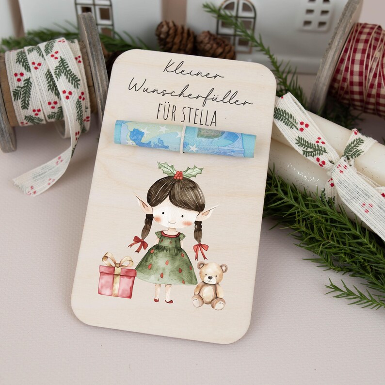 May include: A wooden card with a watercolor illustration of an elf holding a gift and a teddy bear. The card has the text "Kleiner Wunsch erf&uuml;ller f&uuml;r Stella" and a rolled up bill of money.