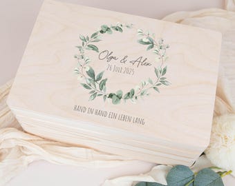 May include: A light-colored wooden box with a hinged lid. The box is decorated with a watercolor wreath of green leaves and white flowers. The text "Olga & Alex 26 July 2025" is centered within the wreath, with the phrase "HAND IN HAND EIN LEBEN LANG" below.