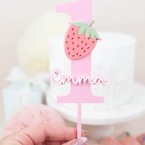 Op de afbeelding: Roze taarttopper in de vorm van het cijfer één, versierd met een aardbei en de naam "Emma". De aardbei heeft rode en groene details. De topper wordt voor een witte taart gehouden.