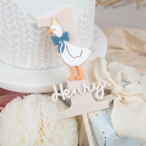 May include: A pink and white cake topper featuring a white duck wearing a blue bow and a yellow hat. The topper is shaped like the number one and has the name "Henry" written on it.