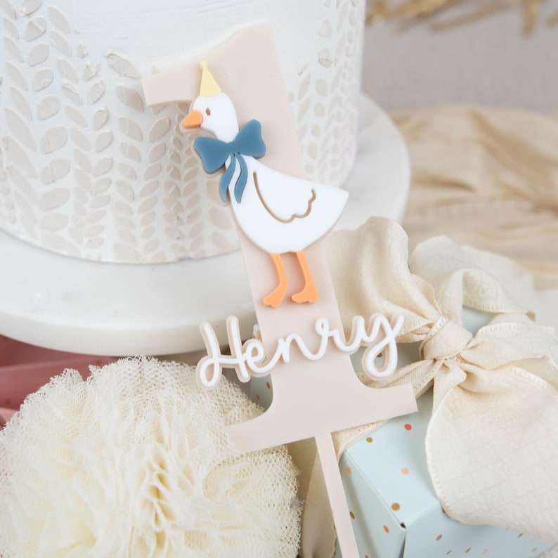May include: A pink and white cake topper featuring a white duck wearing a blue bow and a yellow hat. The topper is shaped like the number one and has the name "Henry" written on it.