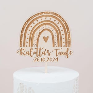 May include: Wooden cake topper featuring a rainbow design with hearts and the text "Kalotta's Taufe 26.10.2024". The topper is made of light-colored wood and is placed on top of a white cake. The rainbow has a heart in the center.