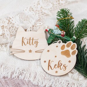 May include: Two wooden Christmas ornaments, one shaped like a cat with the name "Kitty" and the other a round ornament with a paw print and the name "Keks".