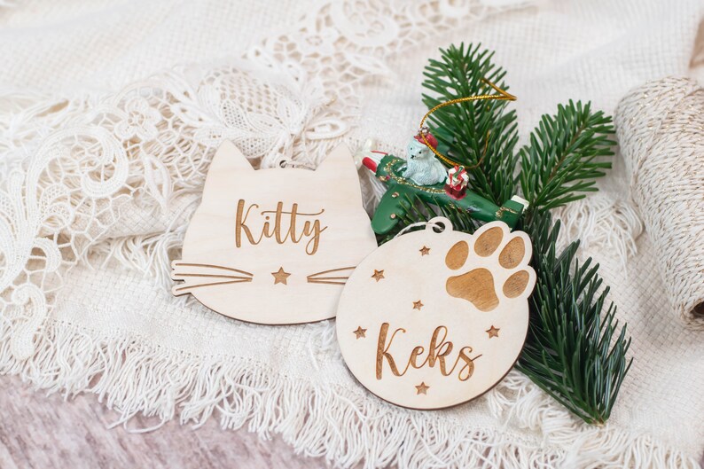 May include: Two wooden Christmas ornaments, one shaped like a cat with the name "Kitty" and the other a round ornament with a paw print and the name "Keks".