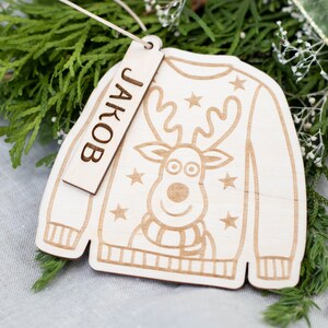 Peut inclure: Une d&eacute;coration de No&euml;l en bois en forme de pull avec un motif de renne et le nom "JAKOB" sur une &eacute;tiquette.
