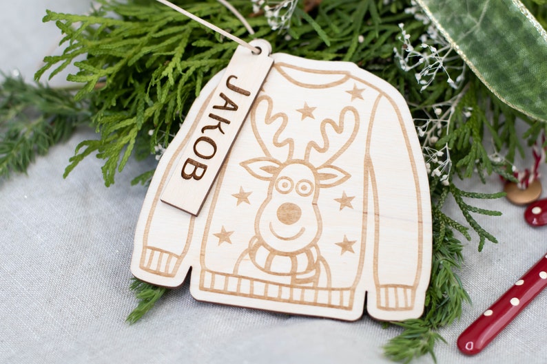 Peut inclure: Une d&eacute;coration de No&euml;l en bois en forme de pull avec un motif de renne et le nom "JAKOB" sur une &eacute;tiquette.