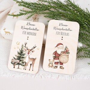 May include: Two wooden Christmas gift tags with illustrations. One tag features a reindeer with a red nose and a Christmas tree with a sign that says "North Pole". The other tag features Santa Claus with a sack of gifts and a teddy bear. The text on the tags reads "Kleiner Wunschert&uuml;ller f&uuml;r Monika" and "Kleiner Wunschert&uuml;ller f&uuml;r Nicolas".