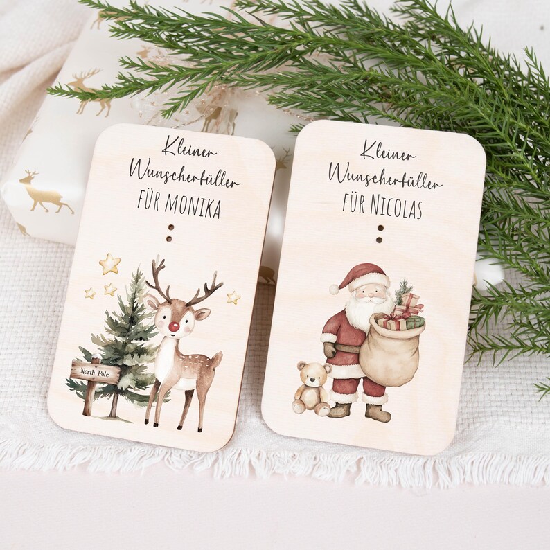 May include: Two wooden Christmas gift tags with illustrations. One tag features a reindeer with a red nose and a Christmas tree with a sign that says "North Pole". The other tag features Santa Claus with a sack of gifts and a teddy bear. The text on the tags reads "Kleiner Wunschert&uuml;ller f&uuml;r Monika" and "Kleiner Wunschert&uuml;ller f&uuml;r Nicolas".