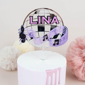 Pode incluir: Um topo de bolo com o nome "LINA" em letras rosa, contra um design de bola de discoteca. O topo inclui notas musicais e uma clave de sol em preto e roxo, montado numa vareta transparente. O bolo é branco com cobertura rosa.