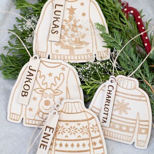 Peut inclure: Quatre d&eacute;corations de No&euml;l en bois en forme de pulls avec des motifs diff&eacute;rents. Chaque d&eacute;coration a une &eacute;tiquette en bois avec un nom dessus : Lukas, Jakob, Charlotte et Evie.