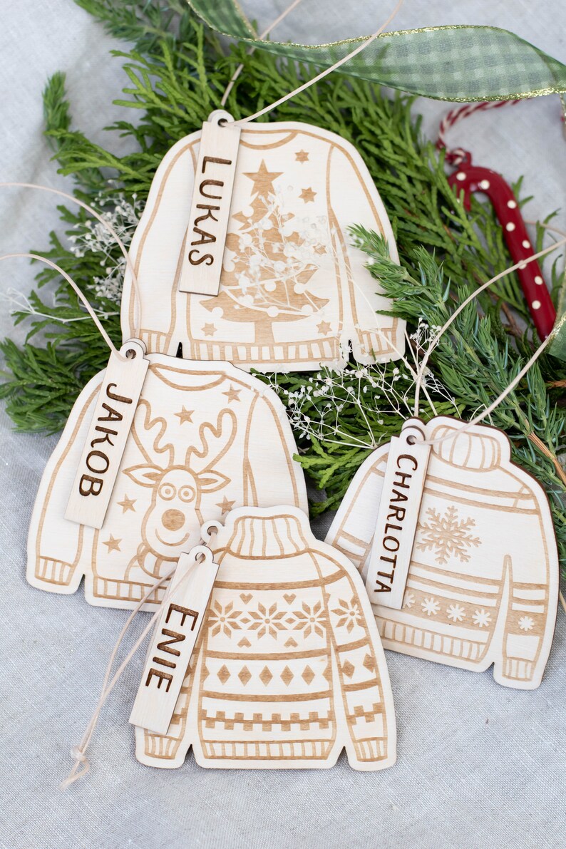 Peut inclure: Quatre d&eacute;corations de No&euml;l en bois en forme de pulls avec des motifs diff&eacute;rents. Chaque d&eacute;coration a une &eacute;tiquette en bois avec un nom dessus : Lukas, Jakob, Charlotte et Evie.