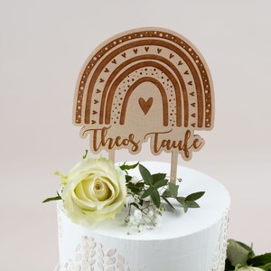 May include: A wooden cake topper featuring a rainbow design with hearts and dots, and the text "Theos Taufe". The topper is placed on a white cake decorated with a white rose and greenery. The cake has a light pink background.