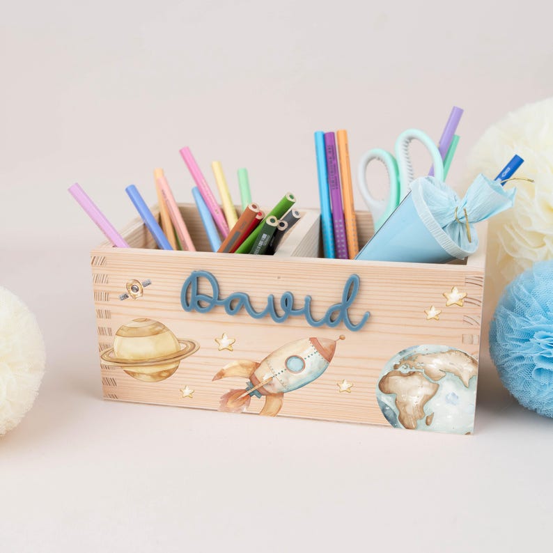 May include: A rectangular wooden desk organiser featuring the name David in blue script lettering. The front is decorated with watercolour illustrations of a planet with rings, a rocket ship, and small stars. It holds various coloured pencils and scissors.