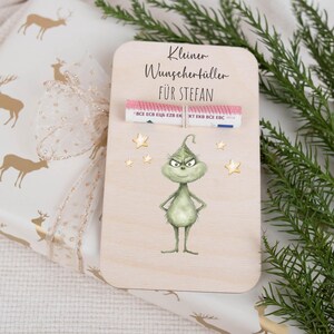 May include: A wooden card with a green Grinch character and gold stars. The card says "Kleiner Wunschert&uuml;ller f&uuml;r Stefan" and has a rolled up bill of money inside.