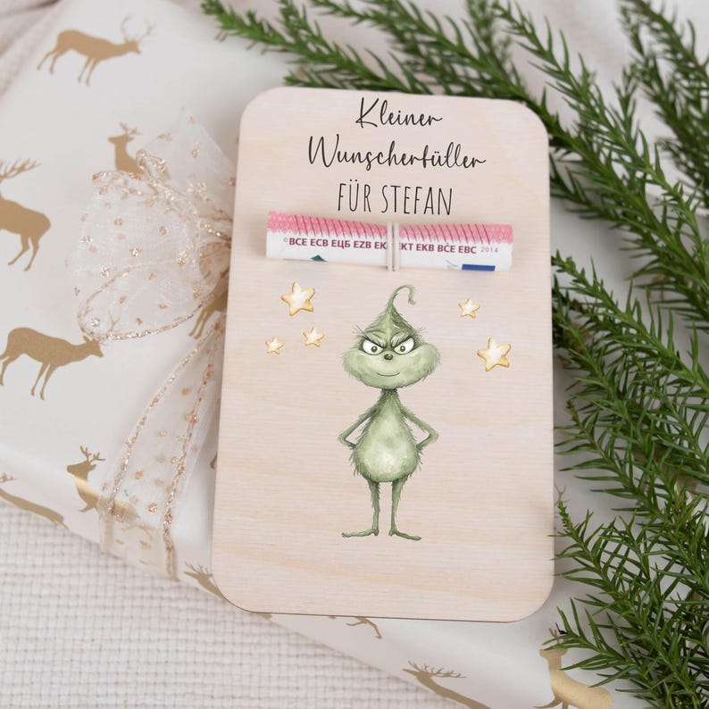 May include: A wooden card with a green Grinch character and gold stars. The card says "Kleiner Wunschert&uuml;ller f&uuml;r Stefan" and has a rolled up bill of money inside.