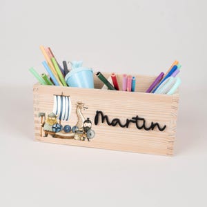May include: A rectangular wooden desk organiser featuring a printed illustration of a Viking ship with a dragon and a warrior. The name Martin is written in black script on the front. The organiser is filled with various coloured pencils and pens.