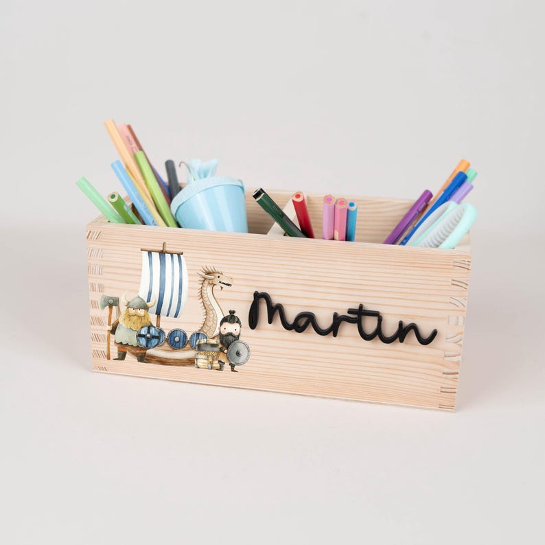 May include: A rectangular wooden desk organiser featuring a printed illustration of a Viking ship with a dragon and a warrior. The name Martin is written in black script on the front. The organiser is filled with various coloured pencils and pens.