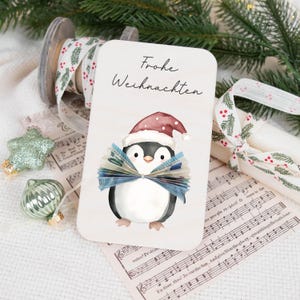 May include: A white card with the text "Frohe Weihnachten" in black lettering. A cartoon penguin wearing a red Santa hat is holding a fan of money in its flippers.