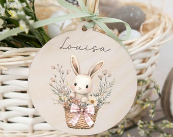 Op de afbeelding: Een ronde houten paasetiket met een konijn in een mandje met bloemen. Het etiket is gepersonaliseerd met de naam "Louisa".