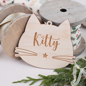 May include: A wooden cat-shaped ornament with the word "Kitty" engraved on it. The ornament is light brown and has a small star in the center of the cat's face.