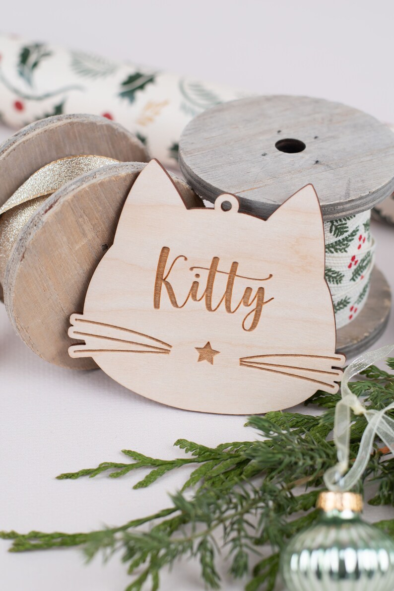 May include: A wooden cat-shaped ornament with the word "Kitty" engraved on it. The ornament is light brown and has a small star in the center of the cat's face.
