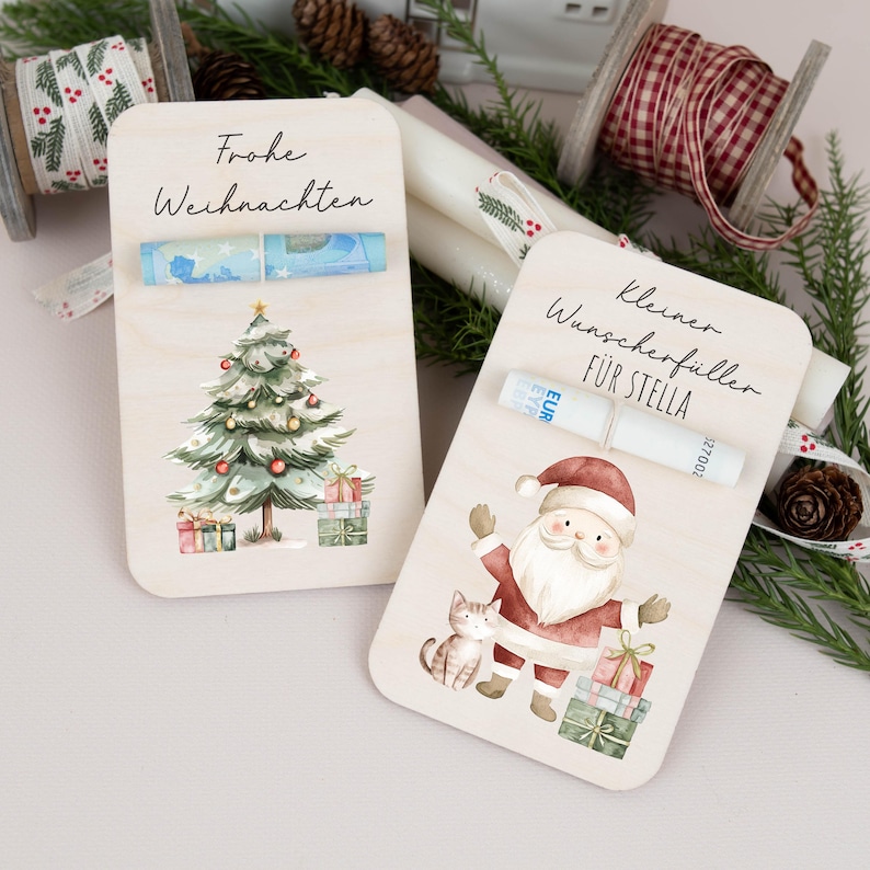 May include: Two wooden Christmas gift cards with illustrations. The first card features a green Christmas tree with presents underneath and the text "Frohe Weihnachten". The second card features a Santa Claus with a cat and the text "Kleiner Wunscherefiller fur Stella".