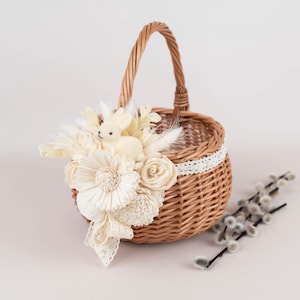 Può includere: Un cestino rotondo in vimini con manico, decorato con fiori color crema, piume e una piccola statuina di coniglietto. Il cestino ha un bordo in pizzo bianco, perfetto per Pasqua o decorazioni primaverili.