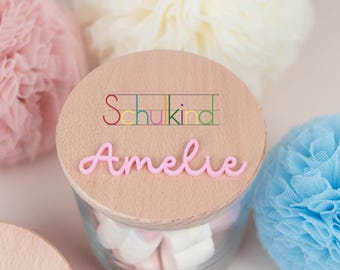 May include: A clear glass jar filled with marshmallows, topped with a wooden lid. The lid features the name "Amelie" in pink script and the word "Schukind" in colorful letters. Decorative pom-poms in pink, white, and blue surround the jar.