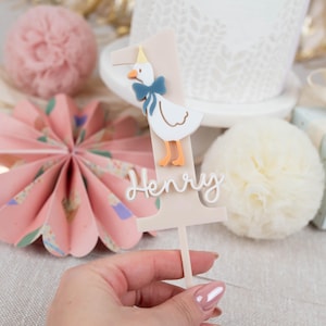 May include: A beige cake topper featuring a white duck wearing a blue bow tie and a yellow hat. The number one is on the topper with the name Henry written below.