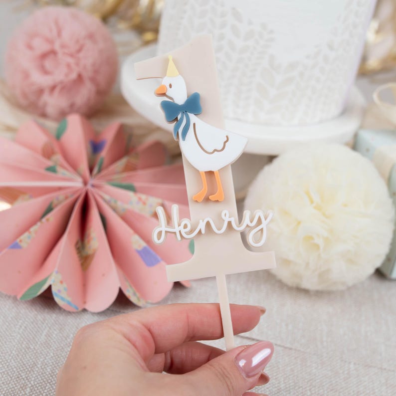 May include: A beige cake topper featuring a white duck wearing a blue bow tie and a yellow hat. The number one is on the topper with the name Henry written below.